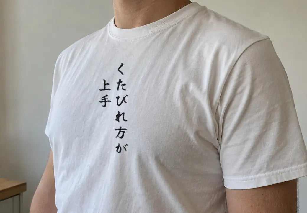 朝の台所で、木のカウンターの横に立つ人。白い T シャツの胸に縦書きで「くたびれ方が上手」（画像生成の人物に、商品と同じ版下を合成）。押すと、白の商品ページを開く