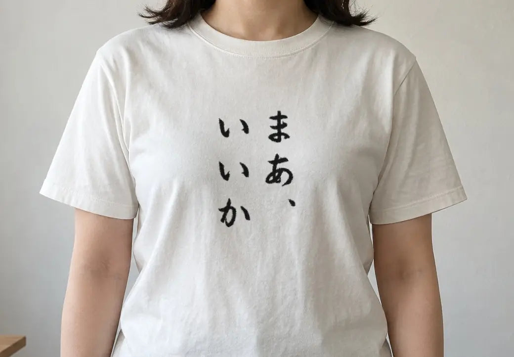 午後の自然光が入る部屋で木のテーブルの横に正面を向いて立つ人。白い T シャツの胸に縦書きで「まあ、いいか」（画像生成の人物に、商品と同じ版下を合成）。押すと、白の商品ページを開く