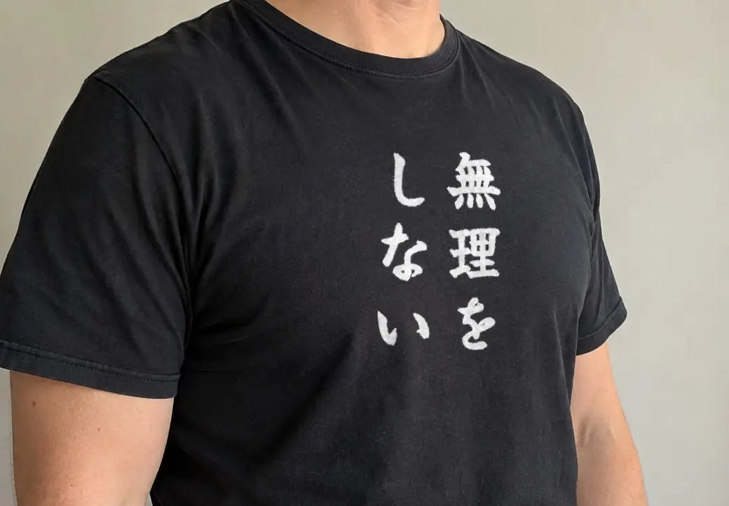 午後、淡い壁の前で体を斜めに向けて立つ人。黒い T シャツの胸に縦書きで「無理をしない」（画像生成の人物に、商品と同じ版下を合成）。押すと、黒の商品ページを開く
