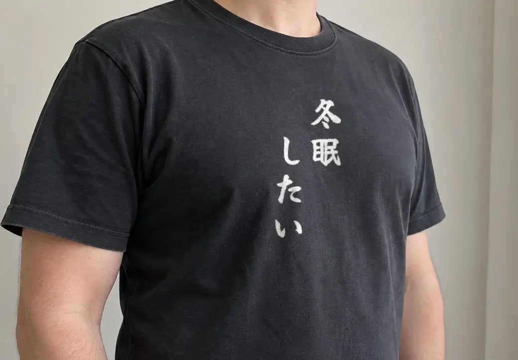 冬のやわらかな光が入るカーテンの横に立つ人。黒い T シャツの胸に縦書きで「冬眠したい」（画像生成の人物に、商品と同じ版下を合成）。押すと、黒の商品ページを開く