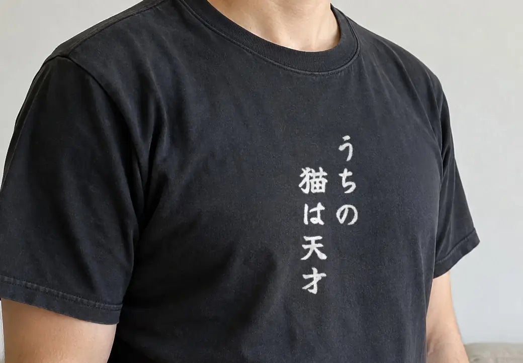 リビングのソファの前で体を斜めに向けて立つ人。黒い T シャツの胸に縦書きで「うちの猫は天才」（画像生成の人物に、商品と同じ版下を合成）。押すと、黒の商品ページを開く